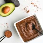 chocolate avocado pudding on a white background with a tablespoon of cocoa powder and halved avocado.
