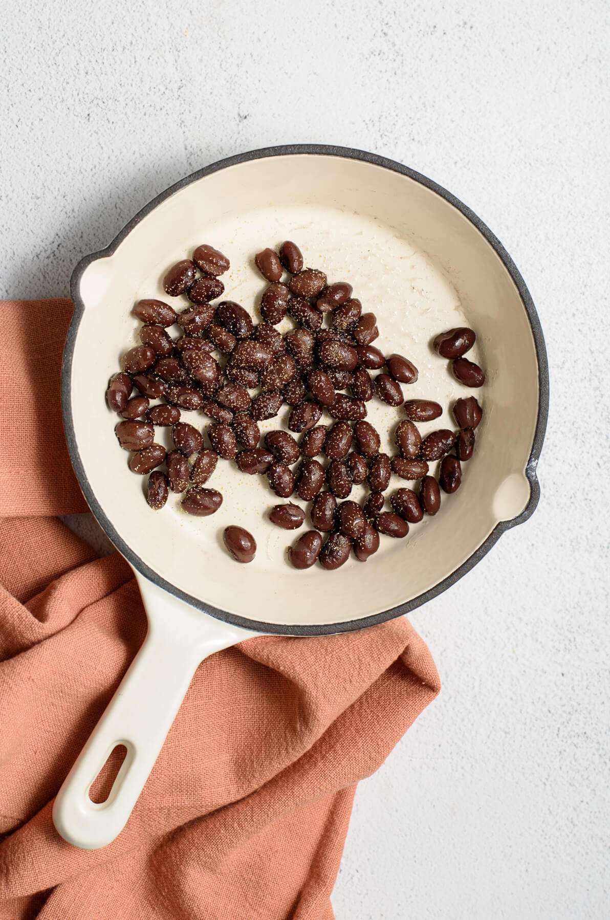 canned black beans cooking on white skillet with seasoning. handle wrapped in pink towel