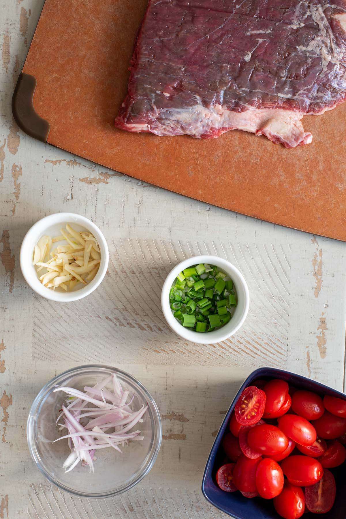 air fryer flank steak ingredients laid out on a table. raw flank steak, a bowl of garlic, scallions, shallots, and tomatoes.