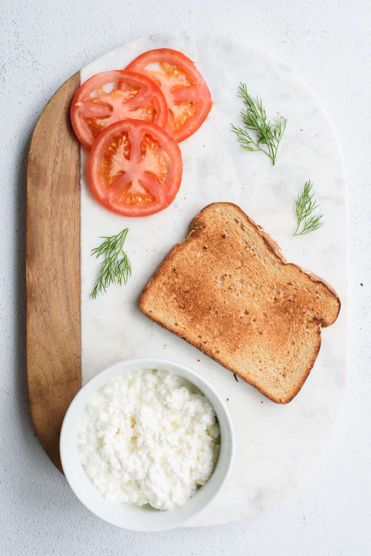 sliced tomatoes laying on cutting board next to one piece of toast, a bowl of cottage cheese, and dill.